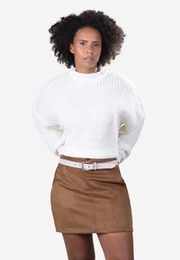 Frau mit lockigem Haar, die einen weißen gestrickten Pullover und einen braunen Wildlederrock mit einem hellrosa Gürtel trägt, steht mit verschränkten Armen hinter dem Rücken.