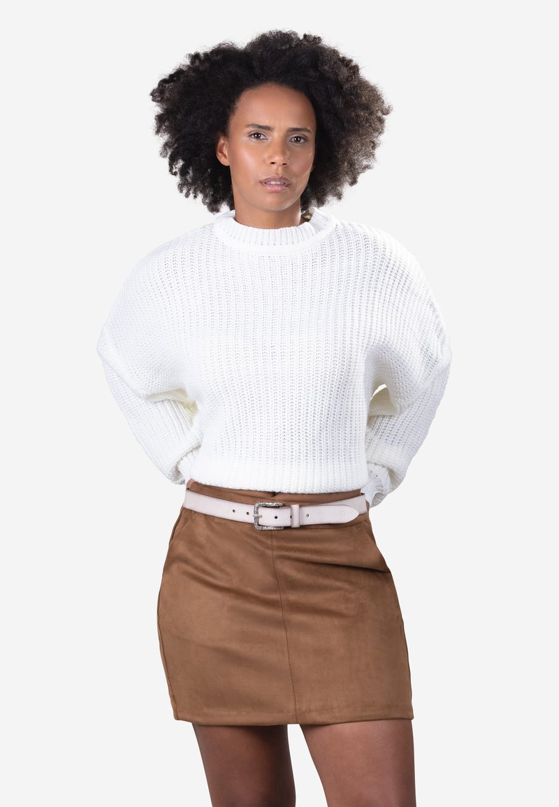 Frau mit lockigem Haar, die einen weißen gestrickten Pullover und einen braunen Wildlederrock mit einem hellrosa Gürtel trägt, steht mit verschränkten Armen hinter dem Rücken.