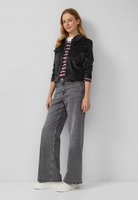 Zwarte spijkerjacket met voorzakken, gecombineerd met een gestreepte langemouwen shirt. Het model draagt een grijs wijde broek en witte sneakers.