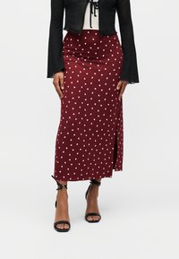 Femme portant une jupe longueur cheville bordeaux avec des motifs de cœurs blancs, un haut noir court et des talons noirs à bride à la cheville.