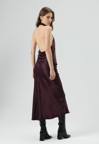 Lange bordeauxrode satijnen jurk met een lage rug, gedrapeerd ontwerp, vloeiende stof en geplooide zijkanten. Gecombineerd met kniehoge zwarte laarzen met hak.