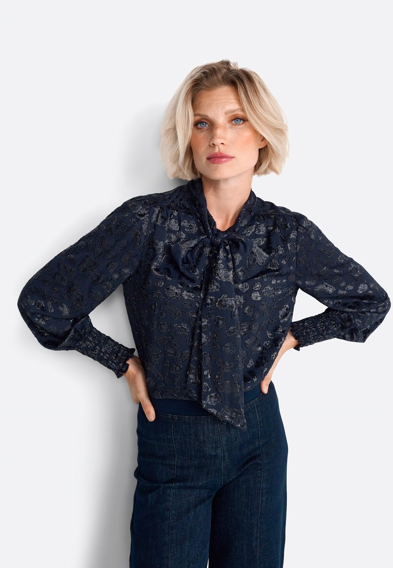 Navy blouse with a textured leopard print, bow tie neck, long sleeves with smocked cuffs, and a fitted hem. Paired with dark denim pants.