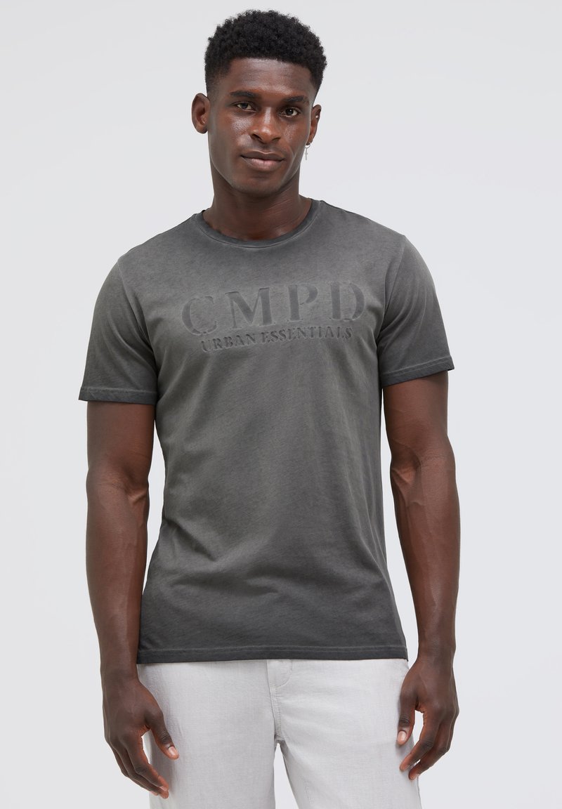 Man wearing a faded black CMPD Urban Essentials t-shirt and light gray pants, standing against a plain light gray background.