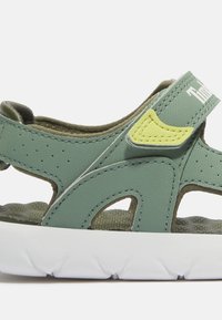 Groene synthetische sandaal met een gestructureerd ontwerp, geperforeerde zijkanten, verstelbare band en een witte rubberen zool. Heeft een geel accentdetail.