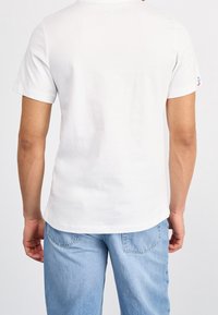 Maglietta bianca a maniche corte con scollo rotondo, realizzata in cotone. Presenta un piccolo logo sulla manica sinistra, abbinata a jeans azzurri.