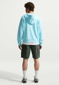 Άνδρας με σγουρά μαλλιά που φοράει ανοιχτό μπλε φούτερ με κουκούλα, λευκή μπλούζα, σκούρα πράσινα σορτς cargo, λευκές κάλτσες και γκρι αθλητικά παπούτσια, στέκεται γυρισμένος από πίσω.