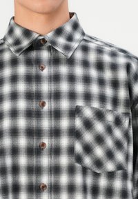 Black and white plaid button-up shirt with a pointed collar, featuring brown buttons and a chest pocket. Textured fabric with contrasting patterns.