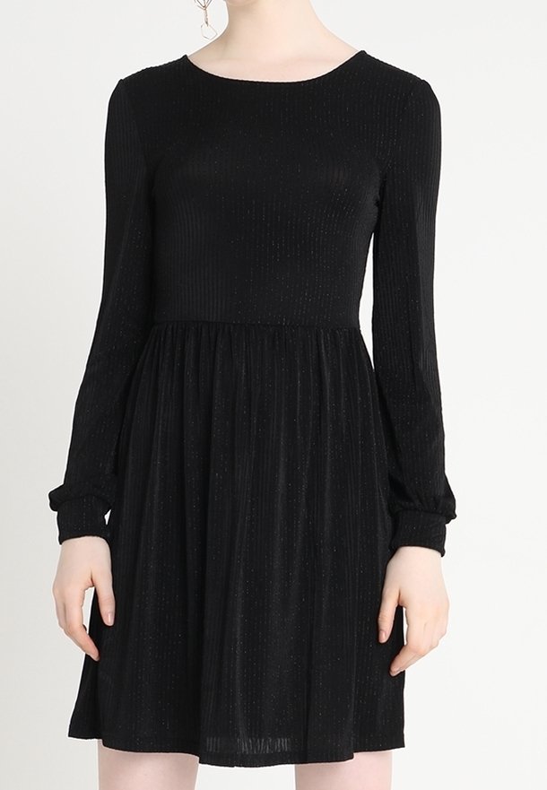 Femme portant une robe noire à manches longues avec un corsage côtelé ajusté et une jupe froncée sur un fond uni.
