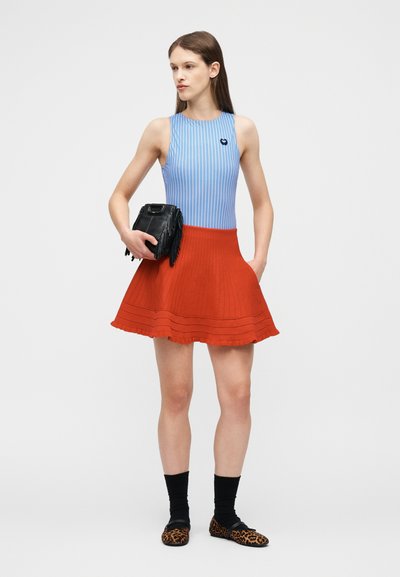 Femme portant un haut sans manches à rayures bleues, une jupe plissée rouge, des chaussettes noires, des chaussures à imprimé léopard, tenant un sac à franges noir.