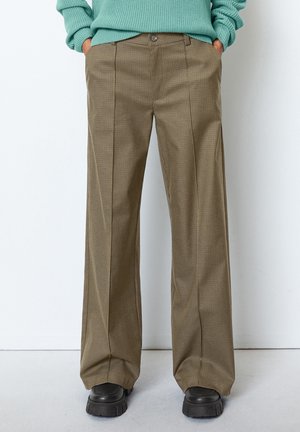 Person iført brune ternede bukser med vide ben, sorte chunky sko og en mintgrøn strikket sweater med hænderne i lommerne.