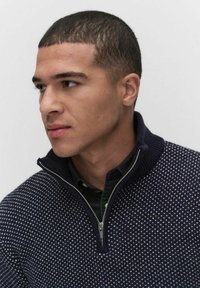 Marinblå stickad tröja med hög krage och halv dragkedja, med ett geometriskt mönster i ljusare blått. Bärs över en mörkgrön skjorta.