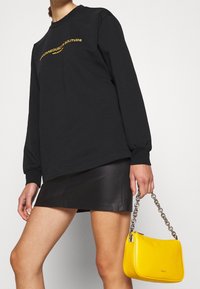Czarna bluza z długim rękawem z napisem "jednocześnie w samotności", zestawiona z czarną skórzaną mini spódnicą oraz żółtą torebką na łańcuszku.