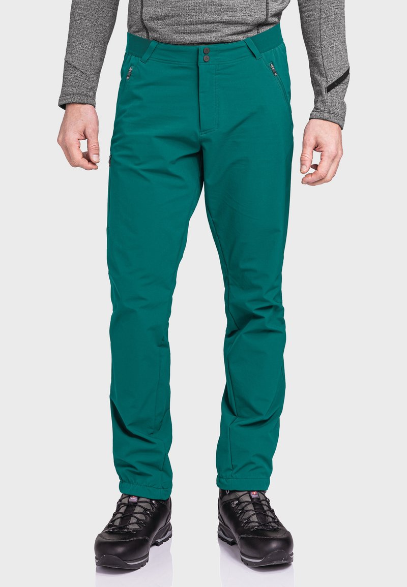 Mann in tealfarbener Outdoorhose mit Reißverschlusstaschen und schwarzen Wanderstiefeln, stehend vor einem weißen Hintergrund.