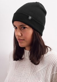 Black knitted beanie with ribbed texture, featuring a logo tag on the front, worn with a white cable-knit sweater.