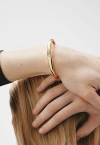 Pulsera de brazalete de metal dorado con un diseño suave y redondeado, y un pequeño acento de piedra clara en el centro, llevada en una muñeca.