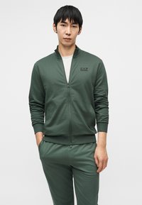 Φούτερ χακί με φερμουάρ, κατασκευασμένο από μαλακό ύφασμα, με πλαϊνές τσέπες και ψηλό γιακά. Το φούτερ συνοδεύεται από τζόγκερς που ταιριάζουν στο ίδιο χρώμα.