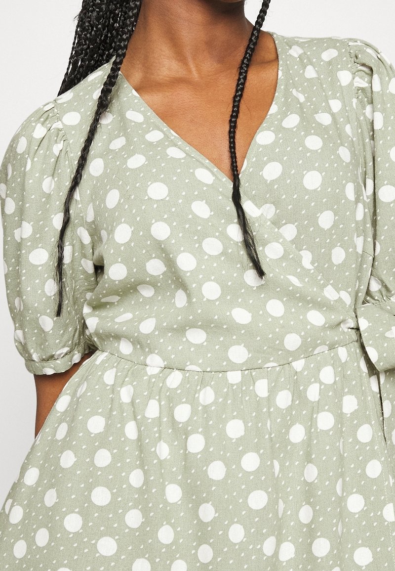 Grön klänning med prickar som har en omvänd design, puffärmar och en samlad midja med en knytdetalj. Tillverkad av en texturerad bomullstyg.