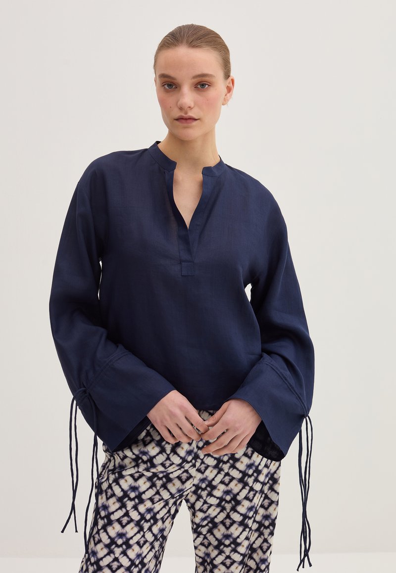 Blusa in lino blu navy con colletto a banda, maniche lunghe e lacci regolabili ai polsini; abbinata a pantaloni stampati.