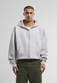 Grauer Reißverschluss-Hoodie mit großer Kapuze, Fronttaschen und gerippten Bündchen. Getragen mit olivgrünen Hosen, die ein entspanntes Fit-Design zeigen.