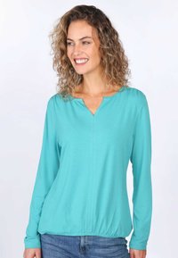 Türkises Langarm-Top mit V-Ausschnitt, weichem Stoff, gerafftem Bund und subtilen Nähdetails. Kombiniert mit blauen Jeans.