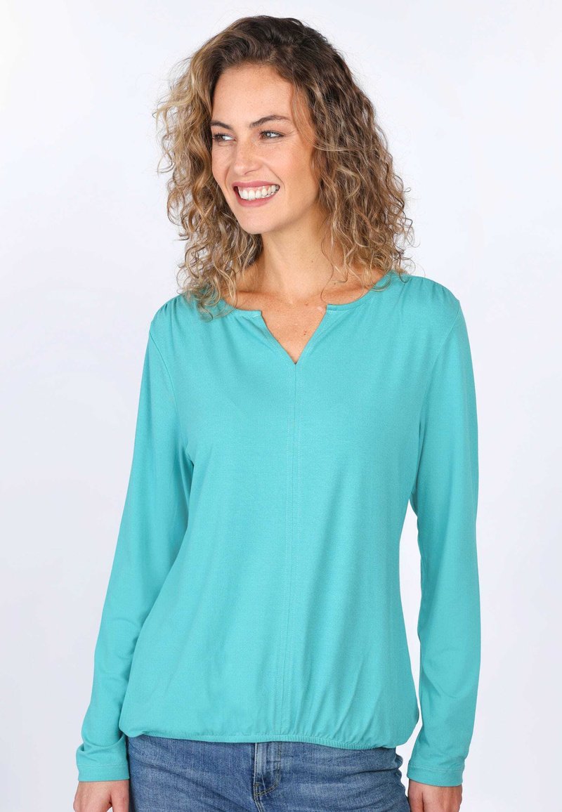 Türkises Langarm-Top mit V-Ausschnitt, weichem Stoff, gerafftem Bund und subtilen Nähdetails. Kombiniert mit blauen Jeans.