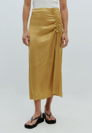 Gelber Satin-Midi-Rock mit gerafften Seitendetails und Bindebändern, kombiniert mit schwarzen Plateausandalen. Glatte Textur und eine taillierte Silhouette.