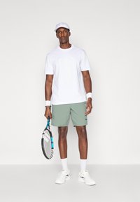 Λευκό T-shirt με σχέδιο λογότυπου, ανοιχτό πράσινο σορτσάκι, λευκό καπέλο, λευκές κάλτσες και λευκά αθλητικά παπούτσια. Κρατά ένα τένις ρακέτα.