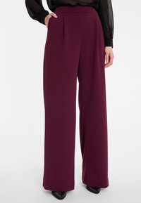 Wijde broekspijpen in een diep bordeauxkleurige stof. Kenmerkt zich door plooien bij de taille en een gladde textuur, gecombineerd met spitse zwarte schoenen.