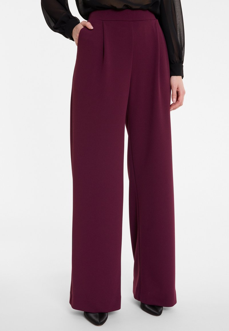 Wijde broekspijpen in een diep bordeauxkleurige stof. Kenmerkt zich door plooien bij de taille en een gladde textuur, gecombineerd met spitse zwarte schoenen.