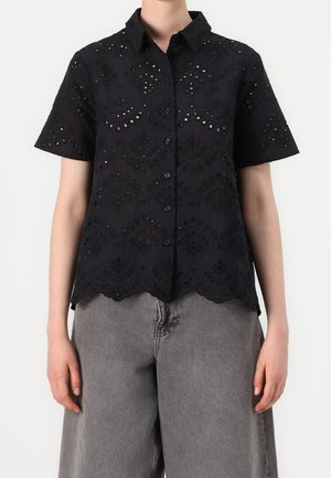Chemise noire à manches courtes avec boutons, broderie ajourée, ourlet festonné, portée avec un pantalon gris ample sur une personne debout devant un fond blanc.