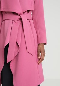 Rosa Wickelmantel mit einem breiten Kragen, langen Ärmeln und einem Stoffgürtel in der Taille. Die Oberfläche wirkt glatt mit einem matten Finish.