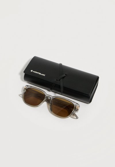 Γυαλιά ηλίου με διάφανο σκελετό και καφετί φακούς δίπλα σε μαύρη θήκη Montblanc που ασφαλίζει με ιμάντα περιμετρικά, πάνω σε ανοιχτό φόντο.