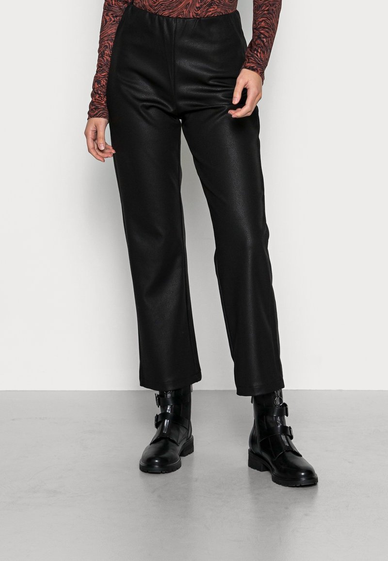 Pantalons en simili cuir noirs à taille haute, avec une coupe droite, texture lisse, et associés à des bottines noires avec détail de boucle.