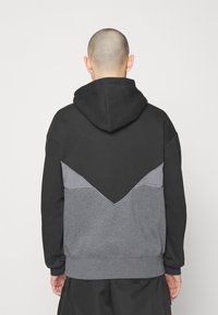 Person med rakad skalle som bär en svart, grå och mörkgrå färgblockerad huvtröja, sedd bakifrån mot en enfärgad bakgrund.