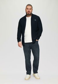 Marineblaues Zip-Up-Jackett über einem weißen Hemd, dunklen Jeans und hellen Turnschuhen. Das Model steht mit neutralem Ausdruck vor einem schlichten Hintergrund.