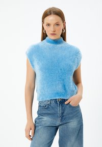 Ljusblå ärmlös topp med hög krage; tillverkad av mjukt, luddigt material. Bärs med ljusblå denimjeans.