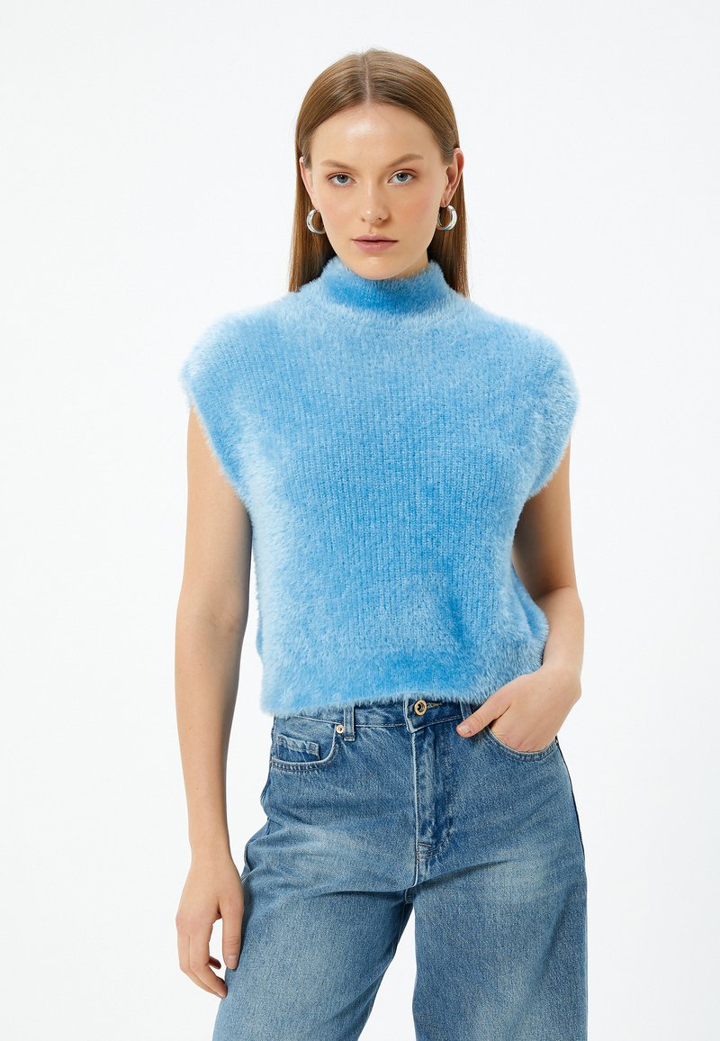 Ljusblå ärmlös topp med hög krage; tillverkad av mjukt, luddigt material. Bärs med ljusblå denimjeans.