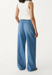 Kvinna klädd i högmidjade vida blå denimbyxor och en vit ärmlös topp, stående framför en enda vit bakgrund.