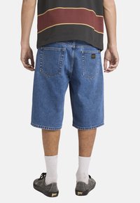 Mann trägt blaue Jeansshorts, weiße Socken und schwarze Turnschuhe, steht mit entspannten Händen an den Seiten vor einem schlichten Hintergrund.