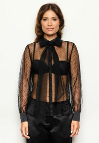 Schwarze, transparente Bluse mit Pünktchenmuster, langen Ärmeln, Schleifenkragen, Knopfverschluss vorne und sichtbarem schwarzen Unterlayer.