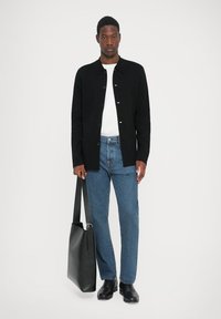 Zwarte button-up jas over een wit T-shirt, gecombineerd met blauwe denim jeans en zwarte schoenen. Houdt een grote zwarte tote bag vast.