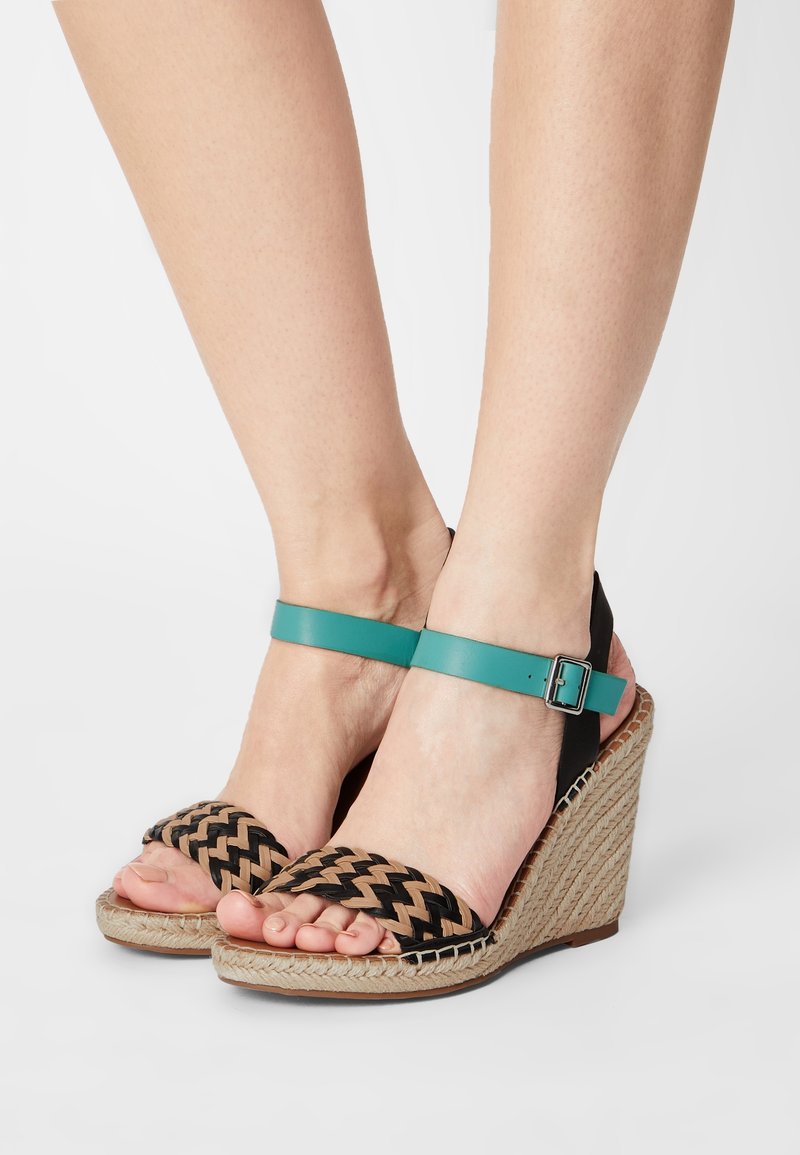 Sandales compensées avec brides avant tressées noires et beiges, brides de cheville bleu sarcelle avec boucles et talons à texture de corde naturelle, portées pieds nus.
