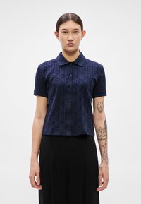 Tumši zila īsām piedurknēm polo krekls ar teksturētu ģeometrisku rakstu un pogām apakšā. Audums šķiet gluds ar strukturētu piegriezumu.