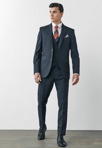 Costum trei piese de culoare gri închis, cu sacou mulat, pantaloni asortați, cămașă albă și cravată cu dungi portocalii și albastre. Pantofi negri cu șireturi completează aspectul.
