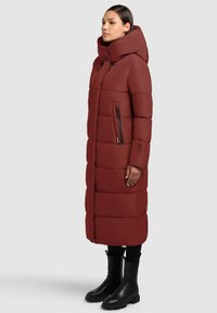 Dlhá červená nadýchaná bunda s vysokým golierom, vybavená vreckami so zipsom, horizontálnym prešívaním a rovnou siluetou. Kombinovaná s čiernymi čižmami.