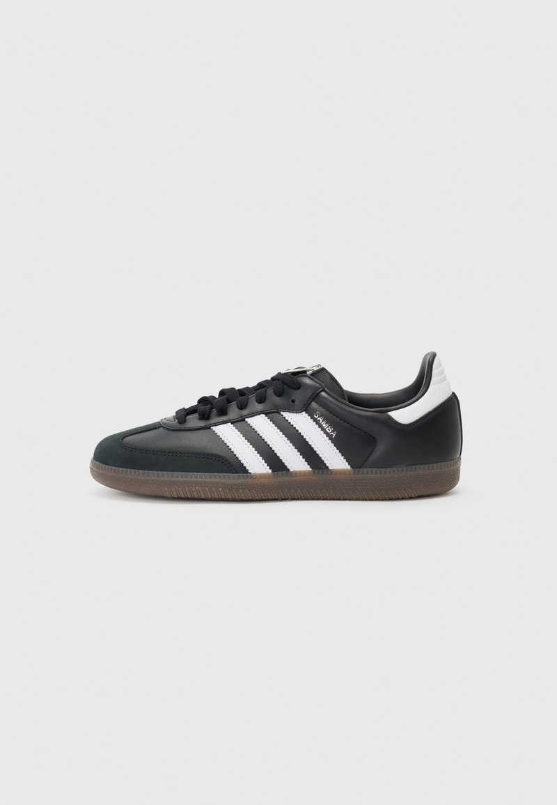 Black synthetic upper sneaker with white three-stripe design, textured grip sole, and black laces. Low-cut athletic style.