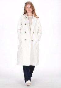 Witte dubbele rij knopen trenchcoat met grote knopen, zijzakken en een licht oversized pasvorm. Gestreept overhemd eronder, donkere spijkerbroek en witte sneakers.