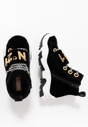 Zwart-witte kindersneakers met gouden veterogen, dikke witte zolen, gestreepte veters, zijritsen en gouden letters op de banden.