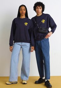 Ναυτικό μπλε φούτερ με κίτρινο γραφικό λογότυπο, σε συνδυασμό με ανοιχτόχρωμα και σκούρα μπλε τζιν παντελόνια. Μαύρη τσάντα κρεμασμένη στον ώμο ενός μοντέλου.