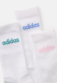 Tri bijele rebraste čarape s Adidas logotipima izvezenim u plavoj, ružičastoj i tirkiznoj boji na rubovima, postavljene preklopljene na bijeloj pozadini.